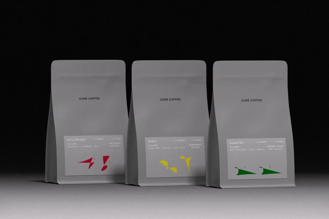 Three grey CORE COFFEE pouches standing in a row with minimalist labels and colorful geometric accents against a dark backdrop.