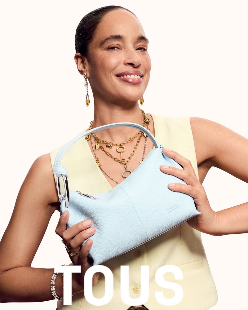 Brandi Quinones smiles while holding a light blue leather Tous handbag against a plain off-white studio background.