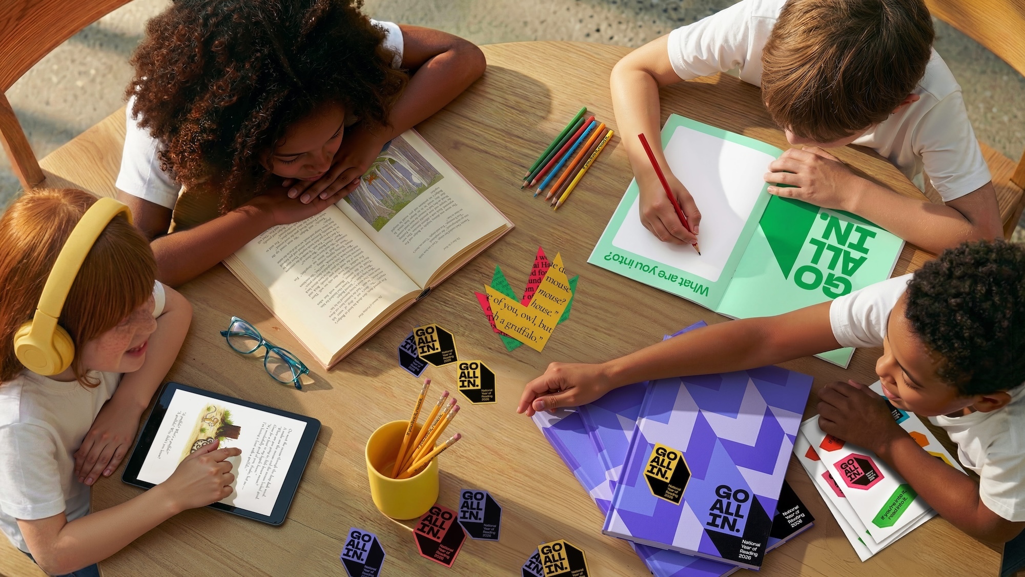 Four children gathered around a wooden table reading books, writing in notebooks, and interacting with tablets.
