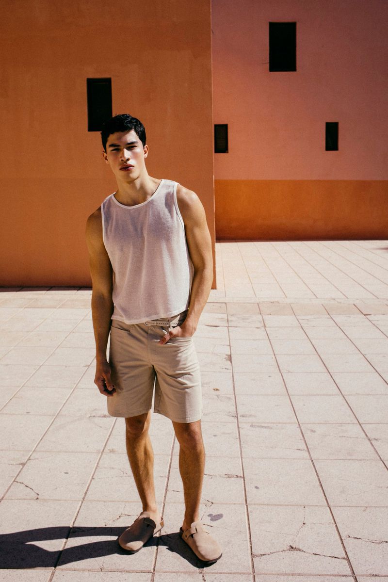 Man standing on a paved outdoor courtyard wearing a white tank top and beige shorts in front of peach terracotta walls.