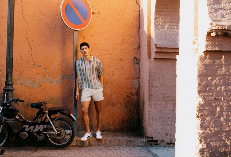 Man in a striped shirt and light shorts leaning on a street pole next to a moped against an orange wall.