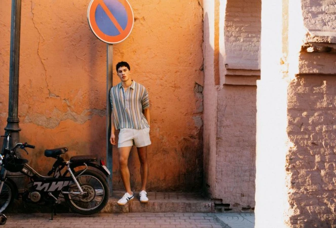 Man in a striped shirt and light shorts leaning on a street pole next to a moped against an orange wall.