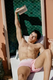Shirtless man in light pink swim shorts lounging on a white sunbed holding a book against green scalloped wall tiles.