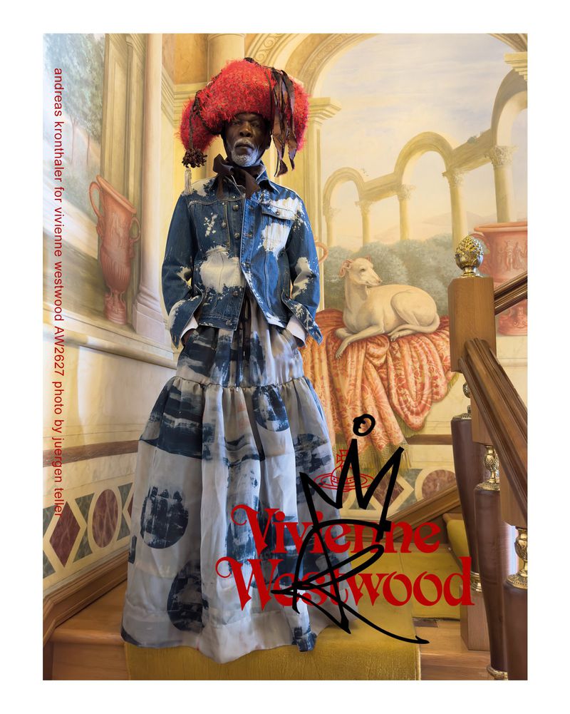Model wearing a blue printed denim jacket, tiered skirt, and oversized red textural hat on a decorated staircase for Vivienne Westwood.