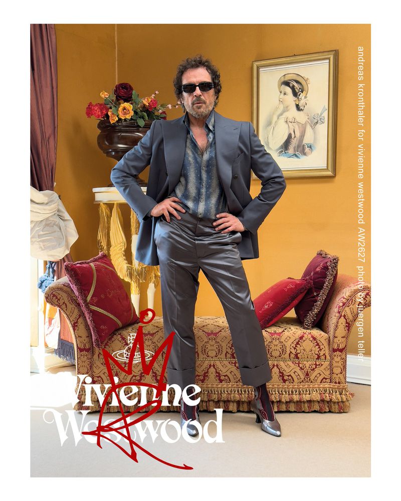 Andreas Kronthaler poses in a grey suit, patterned shirt, and silver heels before an ornate yellow room with a chaise longue.