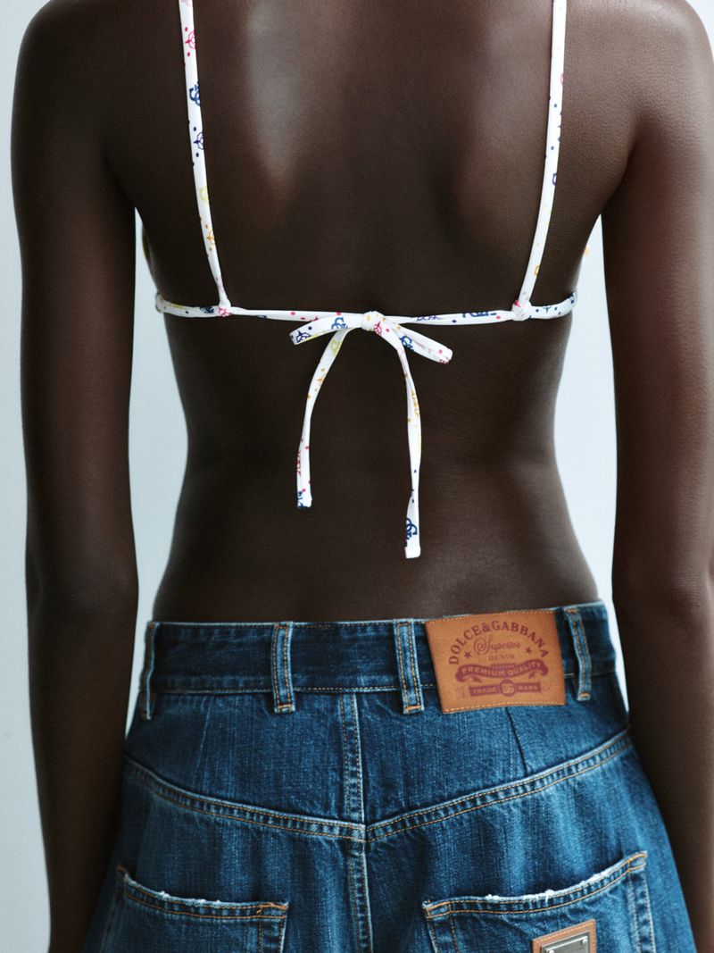 Model seen from behind wearing a white patterned bikini top and blue denim jeans against a white background.
