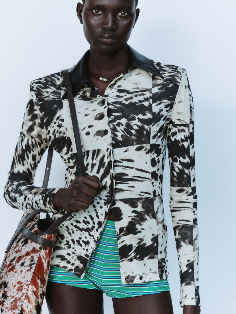 Model Aluel Keror wearing a cow-print long-sleeve shirt with a leather collar and green striped shorts against a plain backdrop.