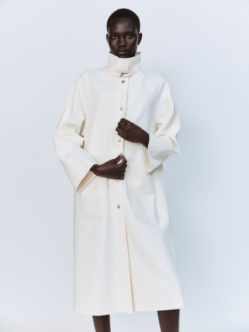 Model Aluel Keror wearing an oversized off-white high-collar buttoned coat against a plain white studio backdrop.