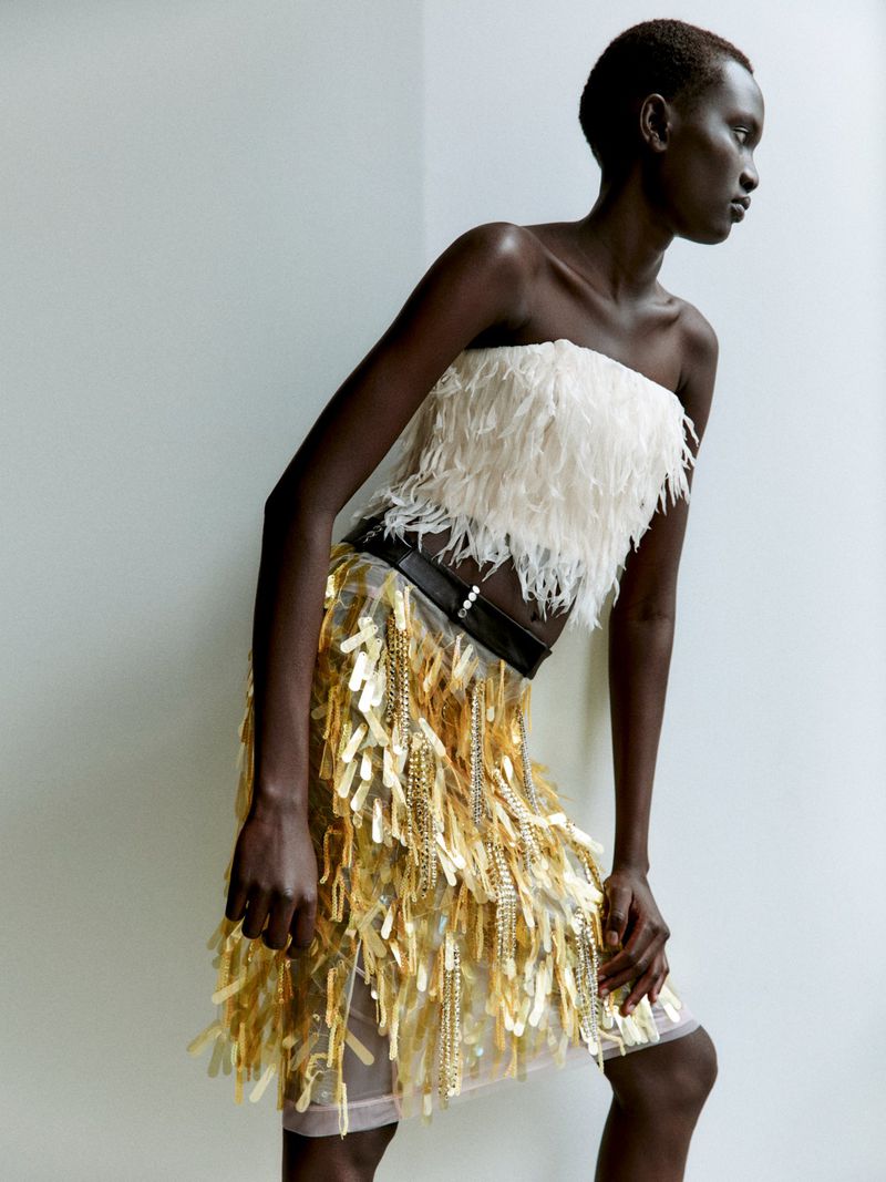 Model Aluel Keror posing in a white feathered strapless top and gold tassel skirt against a neutral studio backdrop.