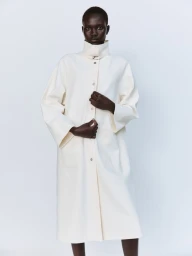 Model Aluel Keror wearing an oversized off-white high-collar buttoned coat against a plain white studio backdrop.