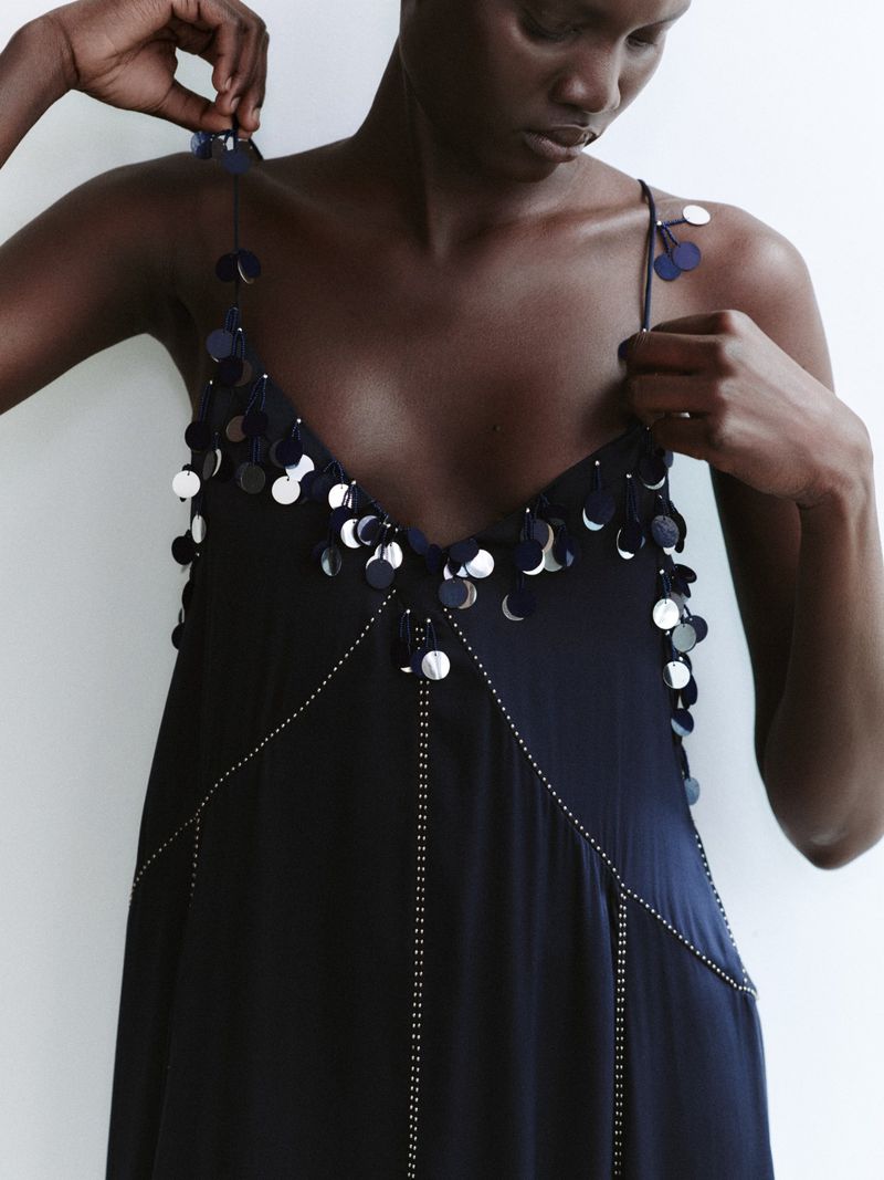 Model Aluel Keror wearing a dark embellished slip dress with circular sequin paillettes against a light studio backdrop.