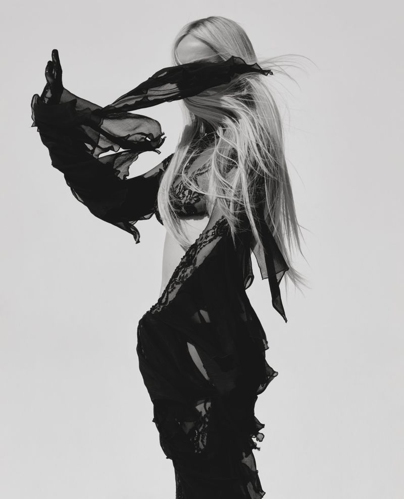 Kim Petras poses in a black ruffled sheer lace ensemble with windblown hair against a white seamless backdrop.