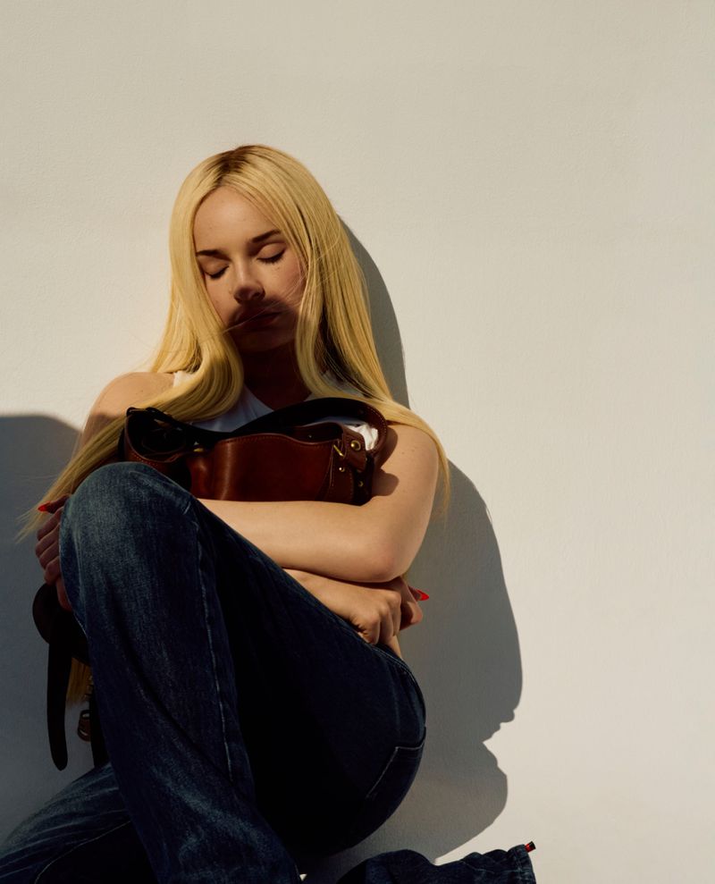 Kim Petras seated with eyes closed, hugging a brown leather shoulder bag in dark denim against a bright white background.