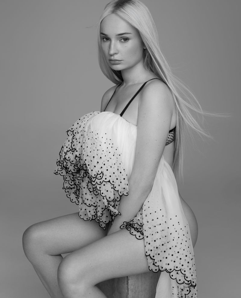 Kim Petras seated in a layered white eyelet ruffle top against a seamless studio background in black and white.