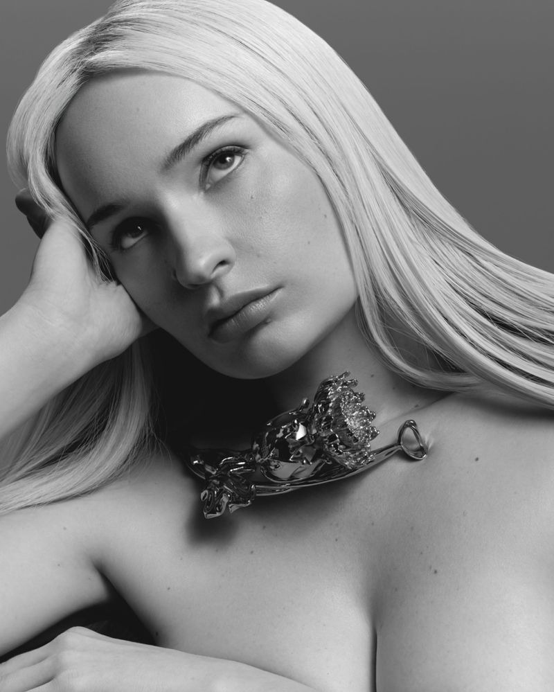 Black and white portrait of Kim Petras with long blonde hair and a sculpted metal flower resting on her collarbone.