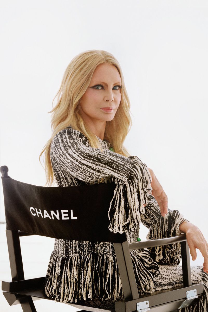 Patty Pravo seated in a black Chanel director's chair wearing a black and white fringed tweed dress against a white studio background.