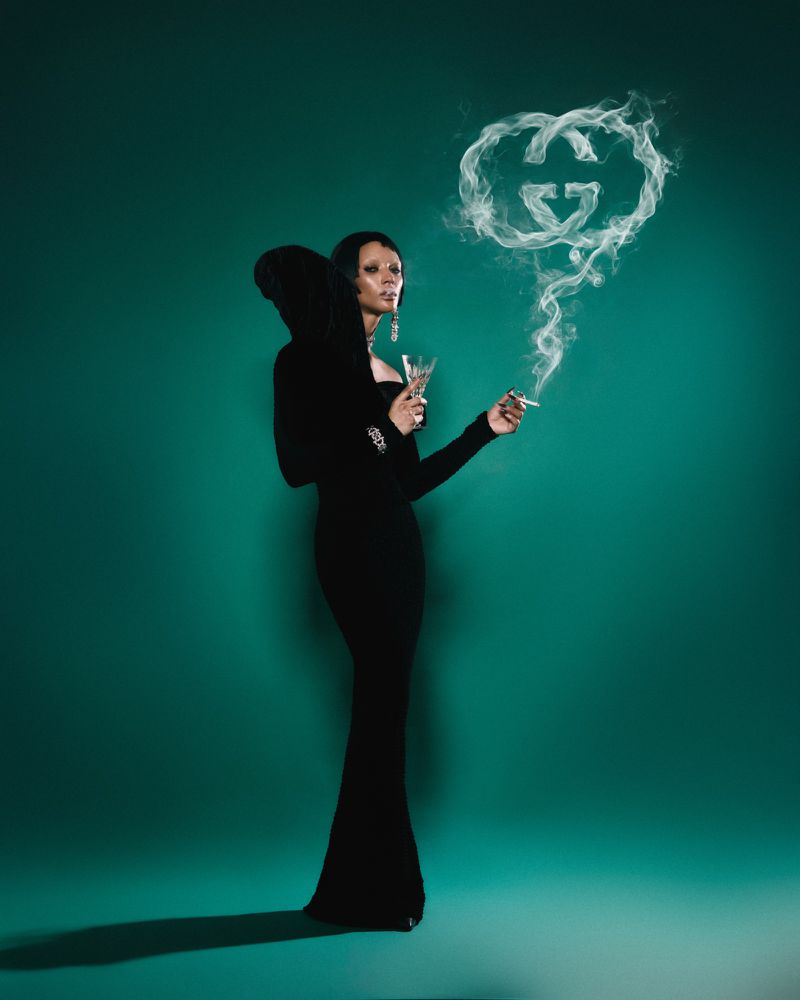 Model Georgia Palmer posing in a black hooded gown holding a cocktail glass and smoking cigarette against a teal studio backdrop.