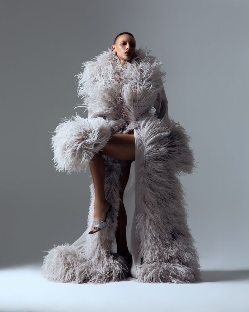 Model Georgia Palmer wearing a voluminous light grey feathered robe and silver heeled mules in a studio setting.
