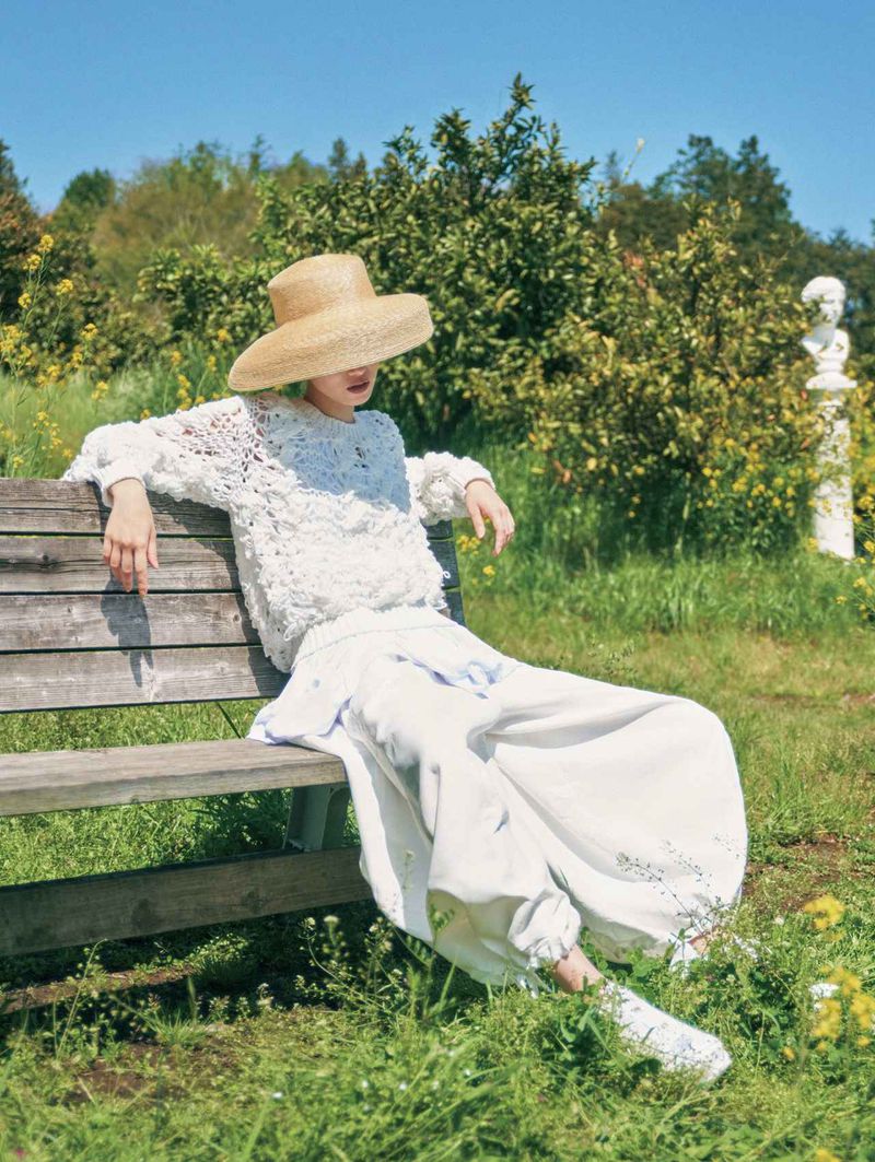 Model Yidan Huang wearing a straw hat and textured white knitwear while seated on a wooden bench in a sunlit garden.