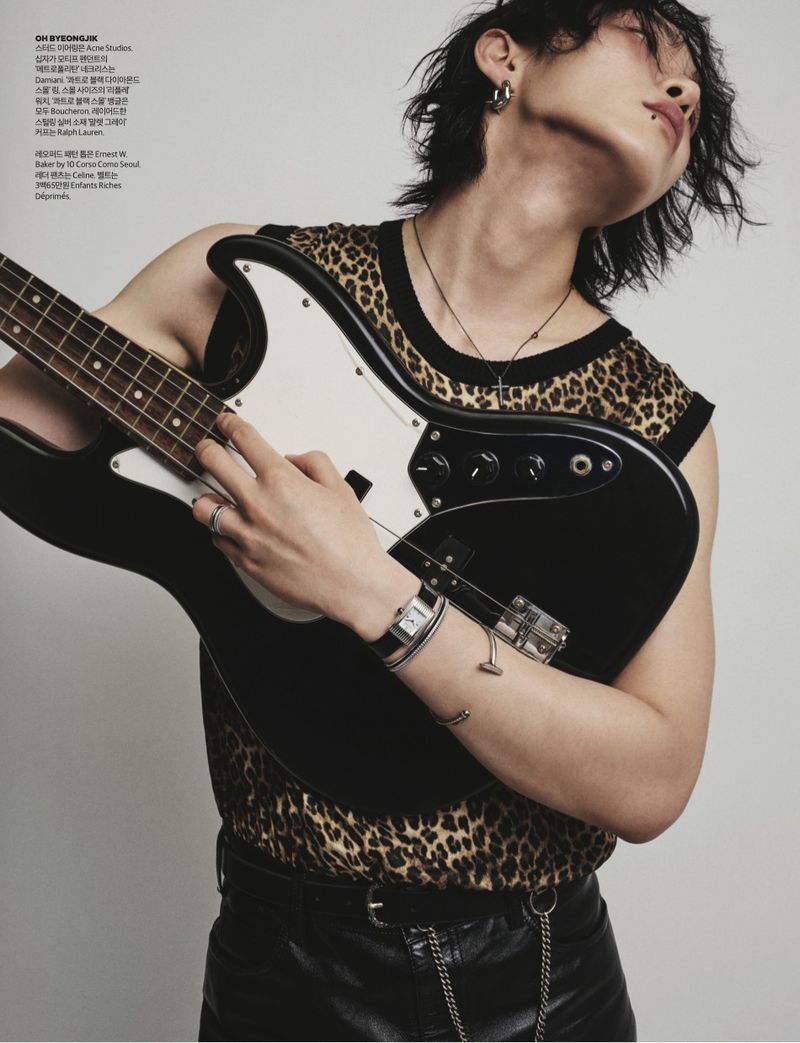 East Asian male model in a leopard print vest holding a black electric bass guitar against a light studio backdrop.