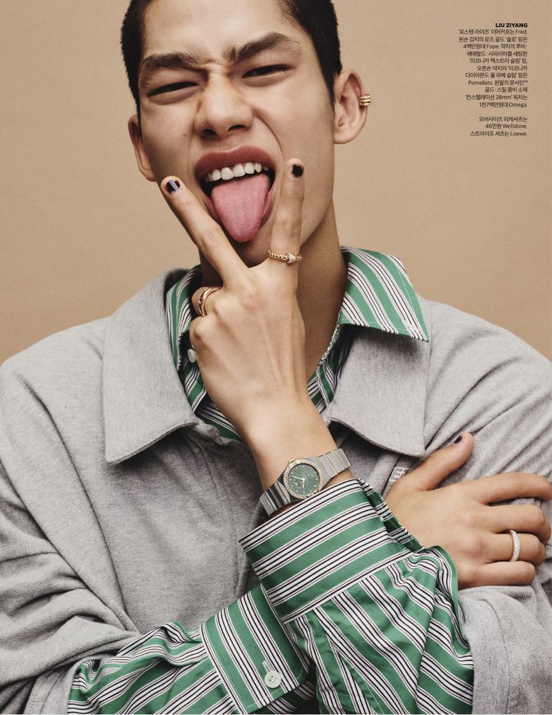 East Asian male model sticking out his tongue wearing a grey jacket, green striped shirt, rings, and a wristwatch against a beige backdrop.
