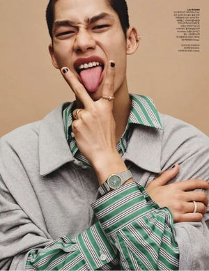 East Asian male model sticking out his tongue wearing a grey jacket, green striped shirt, rings, and a wristwatch against a beige backdrop.