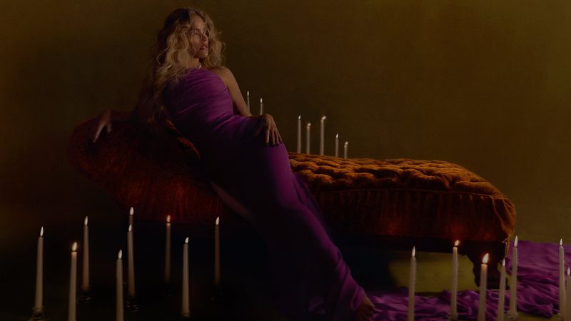 Karol G reclining on a tufted velvet chaise lounge in a purple gown surrounded by lit taper candles.