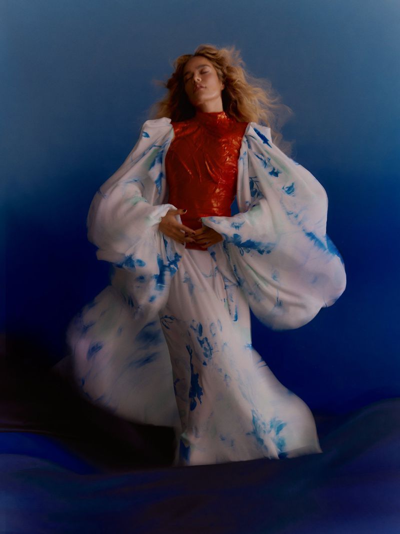 Karol G posing in a voluminous white and blue tie-dye dress with a glossy red high-neck bodice against a blue backdrop.