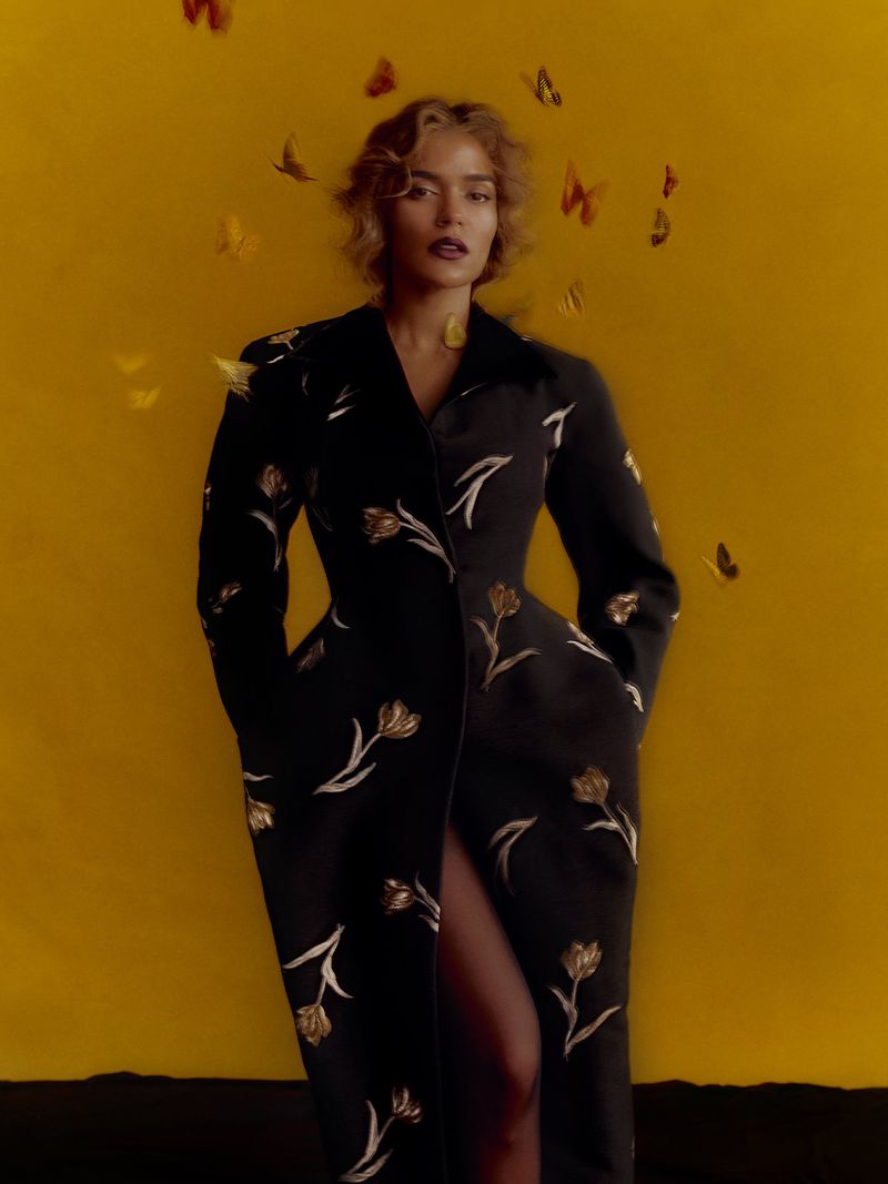 Karol G wearing a black embroidered floral coat surrounded by floating yellow butterflies against a warm yellow backdrop.