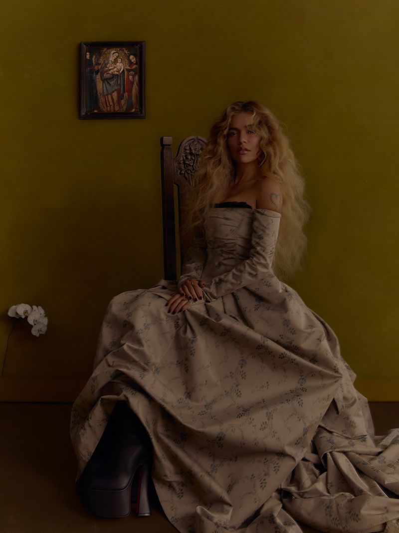Karol G seated in a wooden high-back chair wearing an off-the-shoulder patterned ballgown against an olive green wall.
