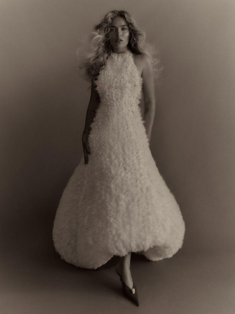 Karol G posing in a voluminous textured halter gown with a bubble hem and pointed heels against a studio backdrop.