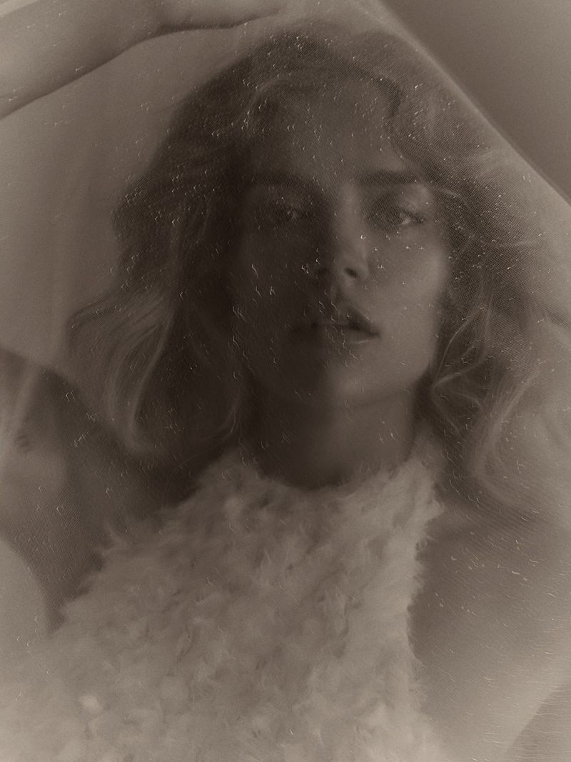 Monochrome portrait of Karol G wearing a textured white halter top seen through a hazy, soft-focus filter.