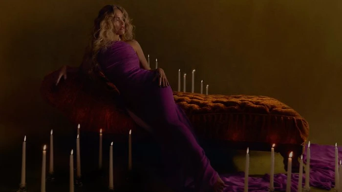 Karol G reclining on a tufted velvet chaise lounge in a purple gown surrounded by lit taper candles.