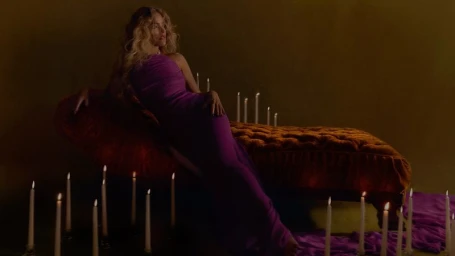 Karol G reclining on a tufted velvet chaise lounge in a purple gown surrounded by lit taper candles.