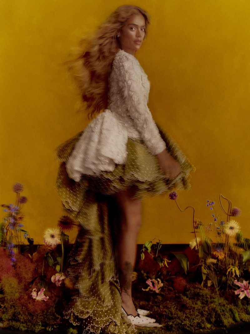 Karol G wearing a white textured top and ruffled olive green skirt standing amid flowers against a yellow backdrop.