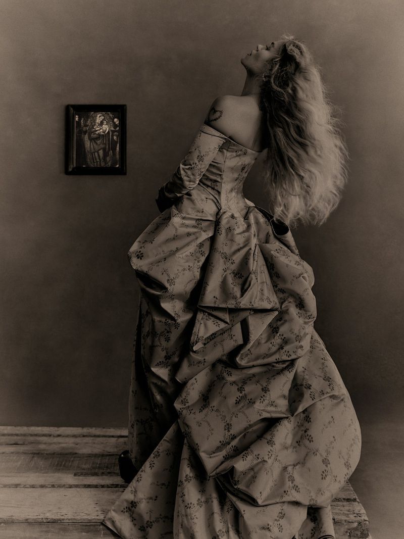 Karol G posing in a voluminous draped brocade ballgown with hair thrown back against a neutral wall with a small framed artwork.