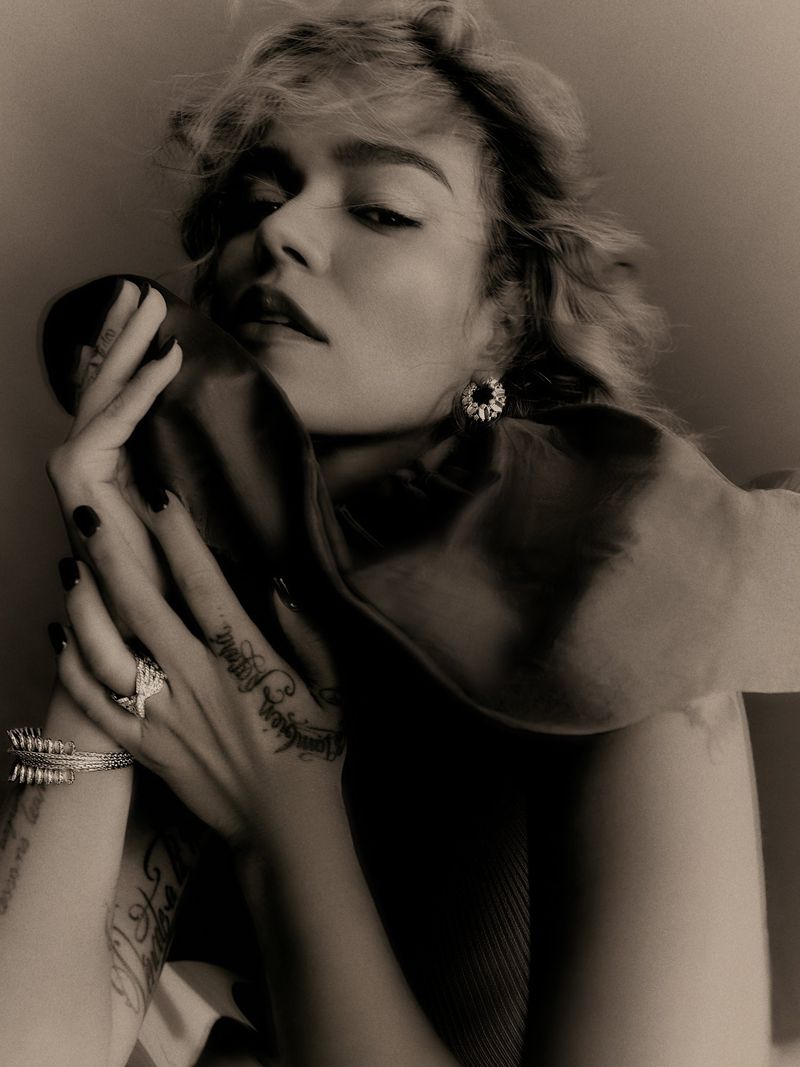 Monochrome close-up portrait of singer Karol G resting her tattooed hands by her face while wearing fine jewellery for Elle U.S.