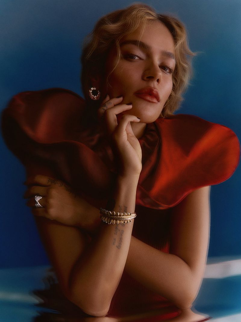 Karol G wearing a sculptural burgundy ruffled garment with gold jewelry against a deep blue studio backdrop.