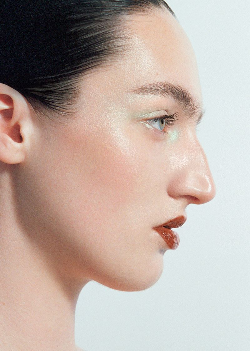 Close-up profile of a woman featuring sleek dark hair, iridescent pastel eye makeup, and glossy brown lipstick against a white backdrop.