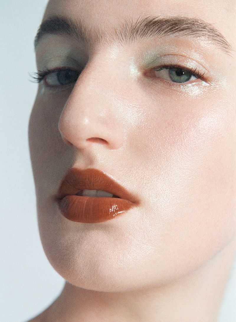 Close-up beauty portrait of a woman with glossy brown lipstick and pale green eyeshadow on a white seamless background.