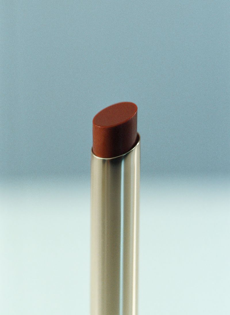 Slanted reddish-brown lipstick in a sleek silver metal tube set against a soft muted blue background.
