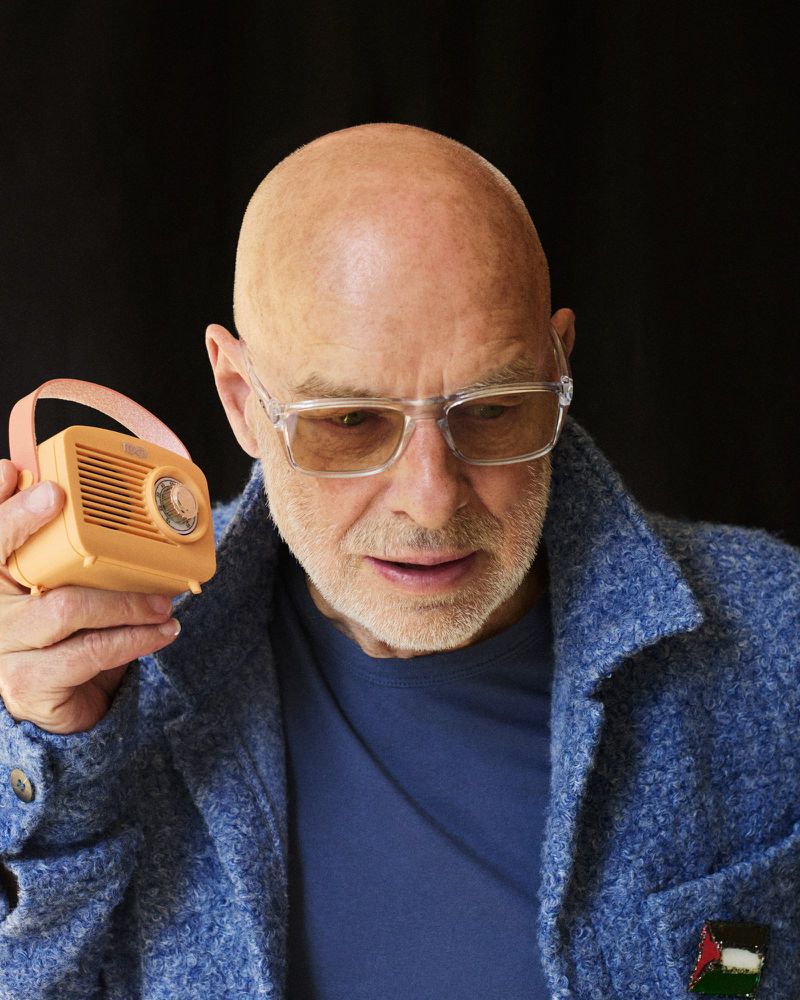 Portrait of Brian Eno holding a small orange portable radio, wearing clear glasses and a blue textured jacket.