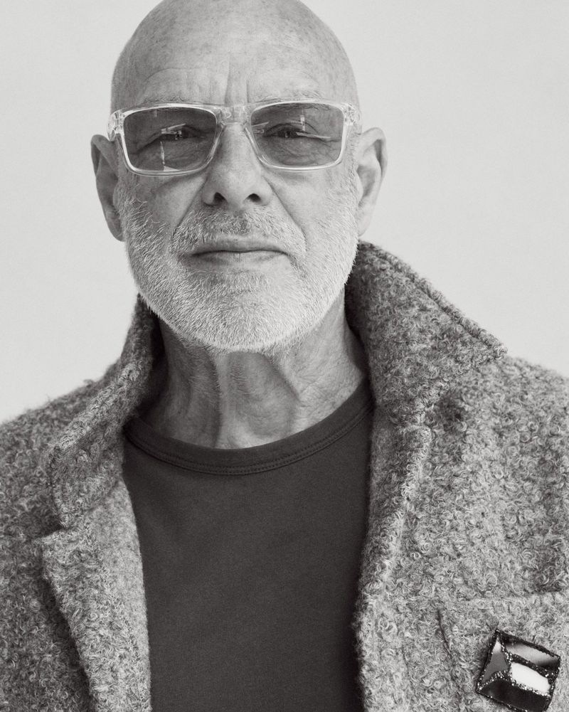 Black and white portrait of Brian Eno wearing clear frame sunglasses and a textured coat against a light studio backdrop.