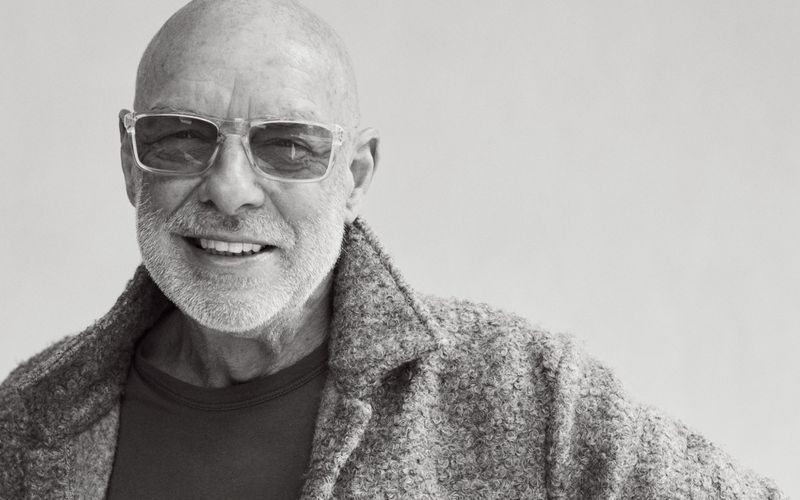Black and white portrait of musician Brian Eno smiling in clear-frame glasses and a textured jacket against a light backdrop.