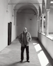 Brian Eno standing with hands in pockets along a sunlit arched cloister in a black and white portrait.