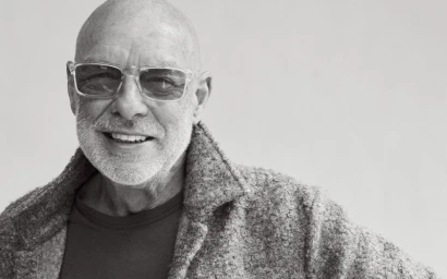Black and white portrait of musician Brian Eno smiling in clear-frame glasses and a textured jacket against a light backdrop.