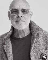Black and white portrait of Brian Eno wearing clear frame sunglasses and a textured coat against a light studio backdrop.