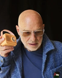 Portrait of Brian Eno holding a small orange portable radio, wearing clear glasses and a blue textured jacket.