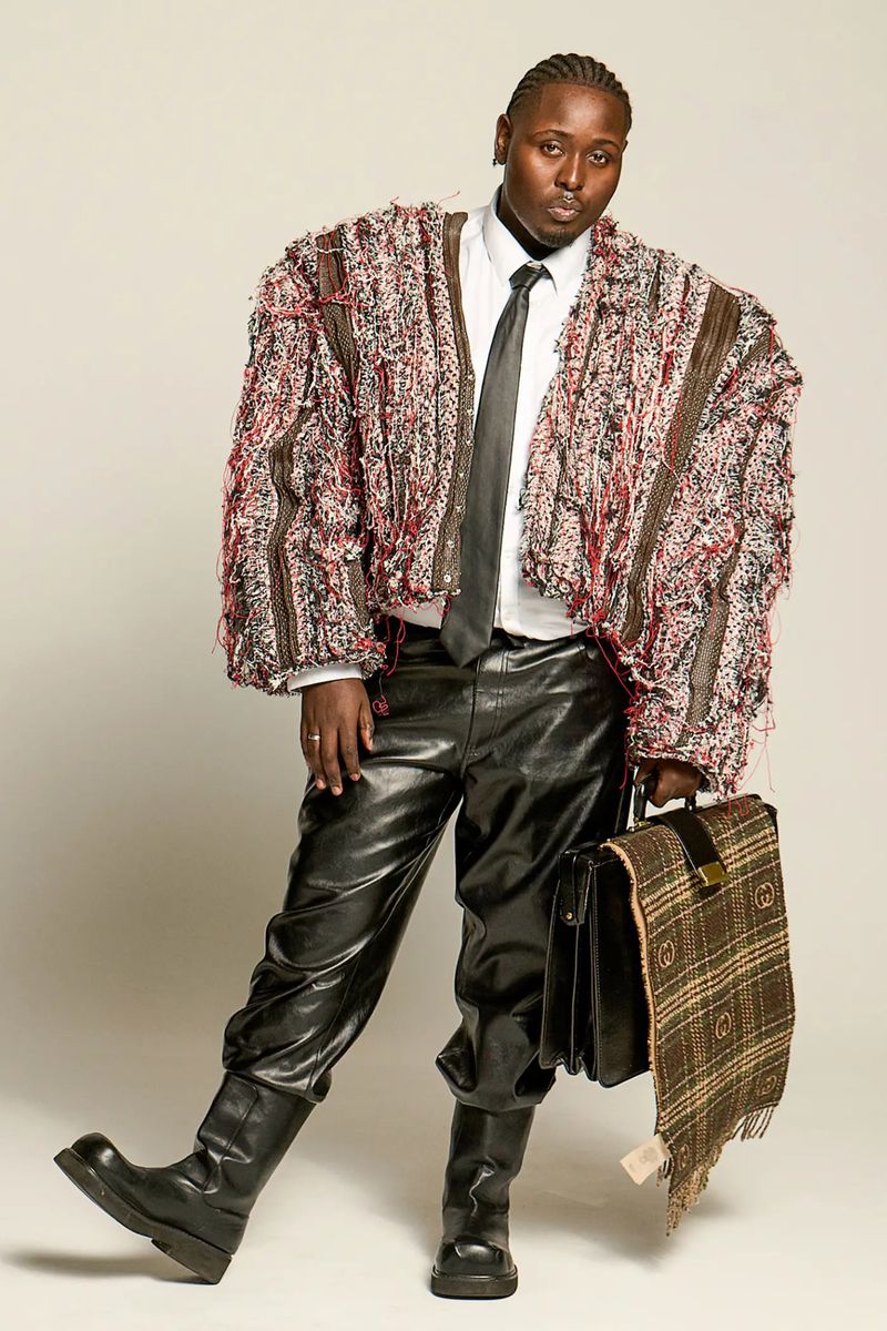 Model James Corbin poses in an oversized fringed tweed jacket, black leather pants, and boots holding a briefcase against a white backdrop.