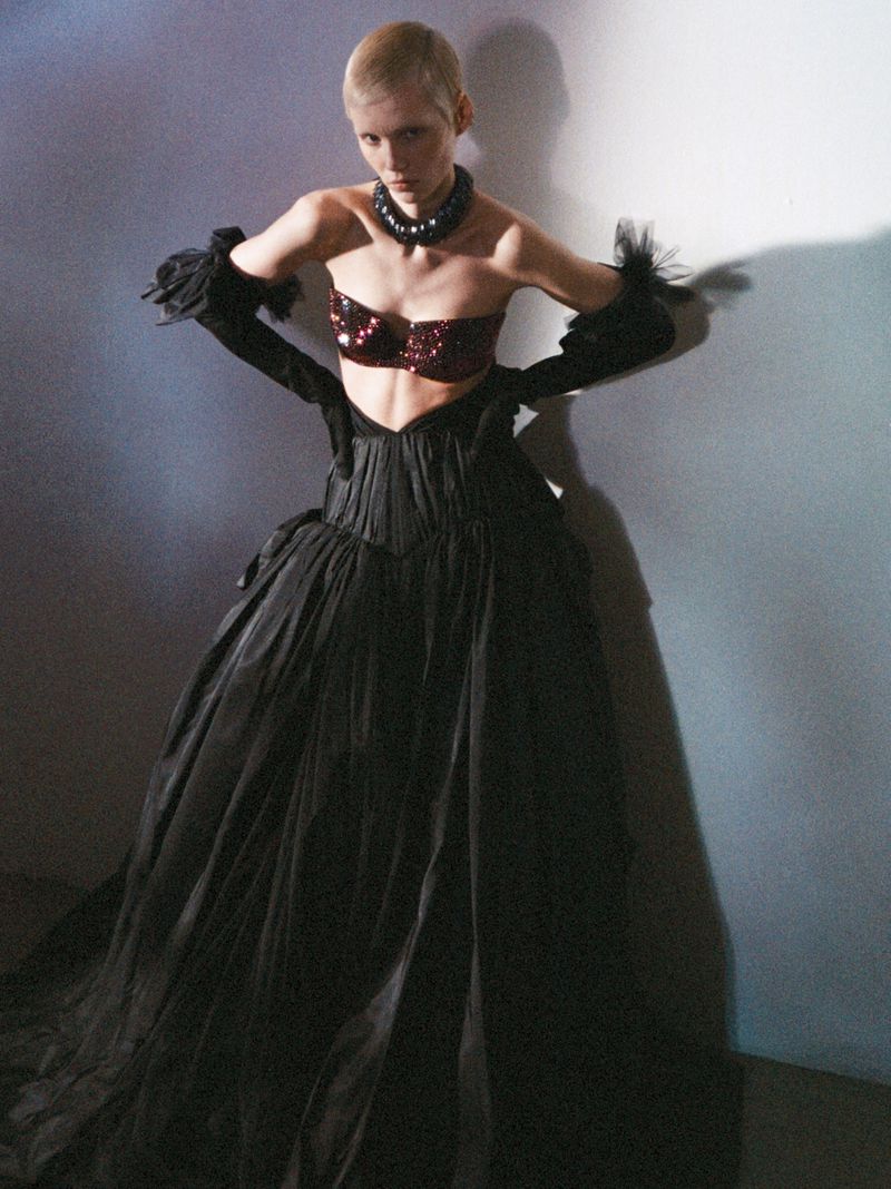Model with short blonde hair in a voluminous black gown, sequined bralette, and long black gloves against a shadowed wall.